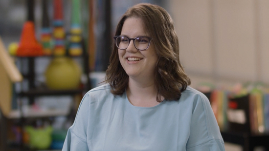 Smiling woman with shoulder-length brown hair and glasses wearing a light blue top, seated indoors with colorful objects on shelves in the background.