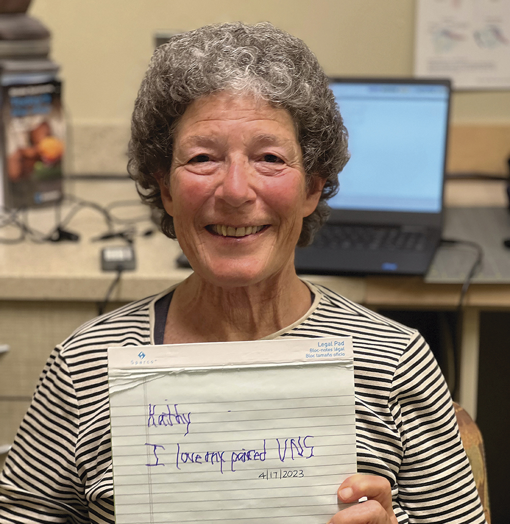 Smiling older woman in a striped shirt holding a handwritten note while sitting in a room with a laptop on the desk in the background.