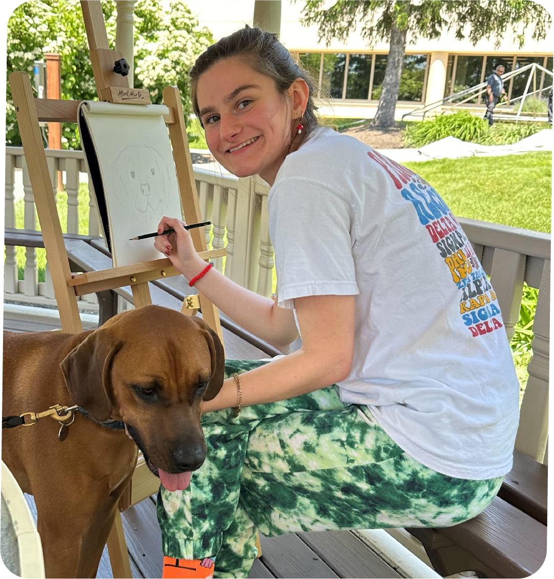 Young woman smiling while drawing on an easel outdoors with a brown dog beside her.